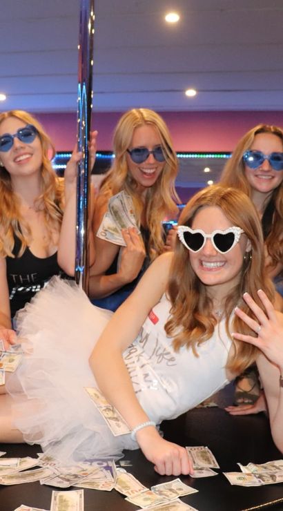 Bachelorette party friends in blue tutus and sunglasses at an indoor pole-party venue, bride-to-be in a white tutu and heart-shaped shades surrounded by dollar bills.