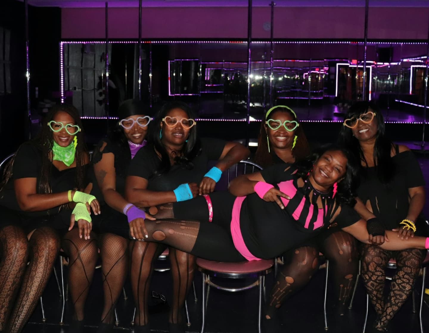 Six women in neon accessories and heart-shaped glasses posing in a purple-lit club/dance studio with mirrored walls and poles, one friend reclining across laps — playful girls' night out group photo