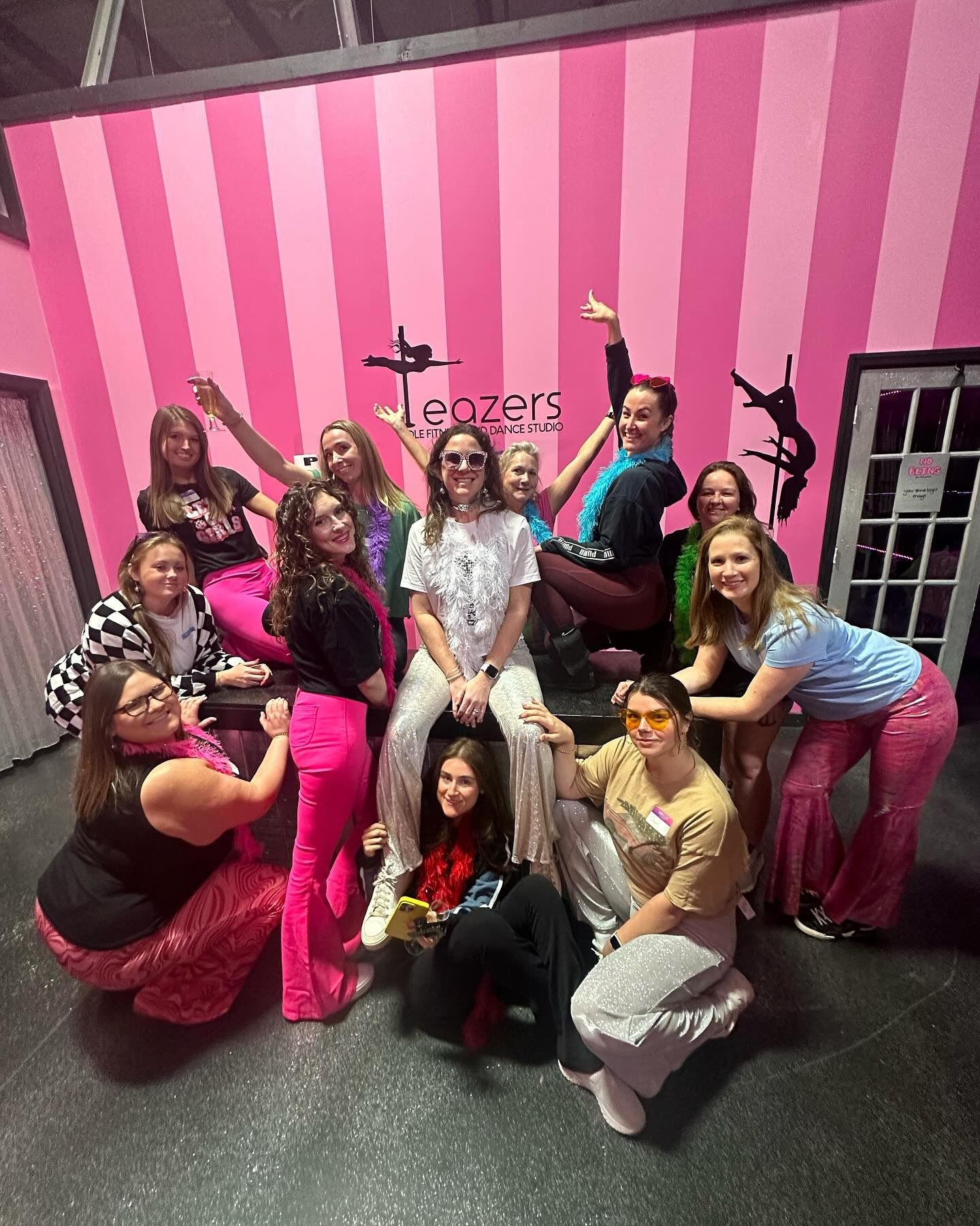 Group of women in colorful retro outfits and feather boas posing and laughing on a bench in a pink-striped pole-fitness studio with pole-silhouette wall art.