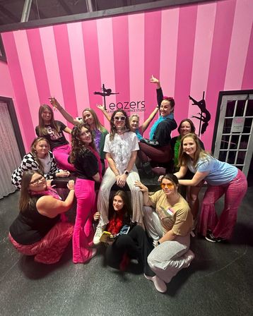 Group of women in colorful retro outfits and feather boas posing and laughing on a bench in a pink-striped pole-fitness studio with pole-silhouette wall art.