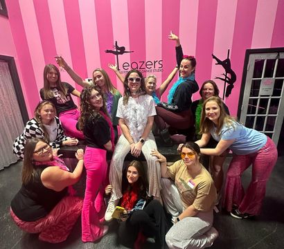 Group of women in colorful retro outfits and feather boas posing and laughing on a bench in a pink-striped pole-fitness studio with pole-silhouette wall art.