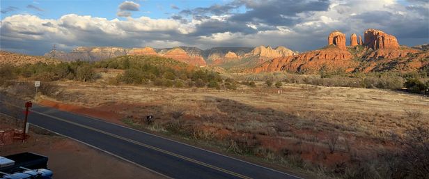 Sunlit Sedona, Arizona red-rock mesas rising above a dry grassy plain with a two-lane road and mailbox in the foreground under dramatic clouds