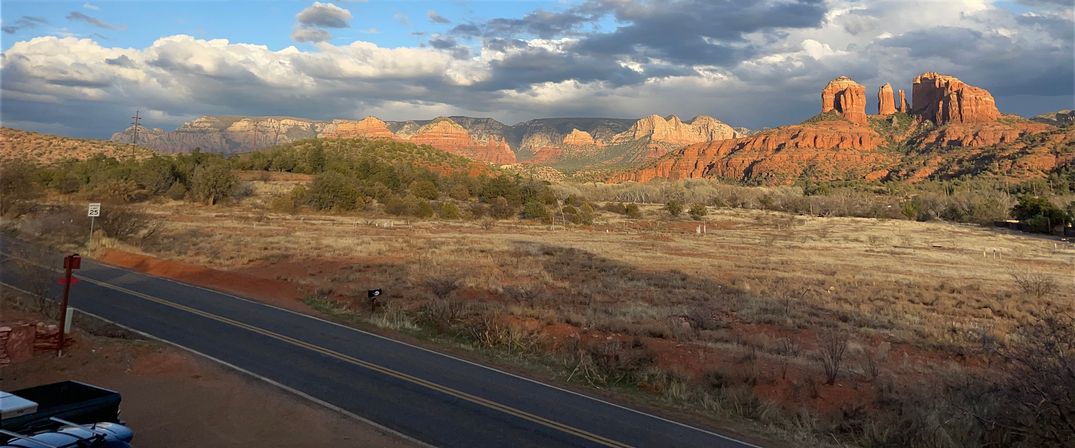 Sunlit Sedona, Arizona red-rock mesas rising above a dry grassy plain with a two-lane road and mailbox in the foreground under dramatic clouds