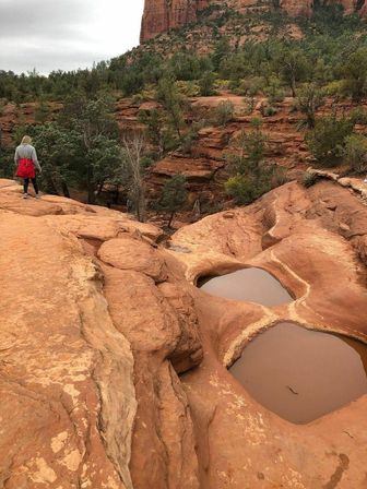 Hiker in a red jacket walks along red sandstone cliffs with rain-filled pothole pools, juniper trees, and towering canyon walls in a Southwestern red-rock landscape.