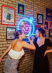 Two friends toast espresso martinis under a blue-and-pink neon record sign on a brick wall lined with vintage music posters in a trendy neon-lit bar.