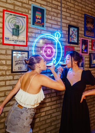 Two friends toast espresso martinis under a blue-and-pink neon record sign on a brick wall lined with vintage music posters in a trendy neon-lit bar.