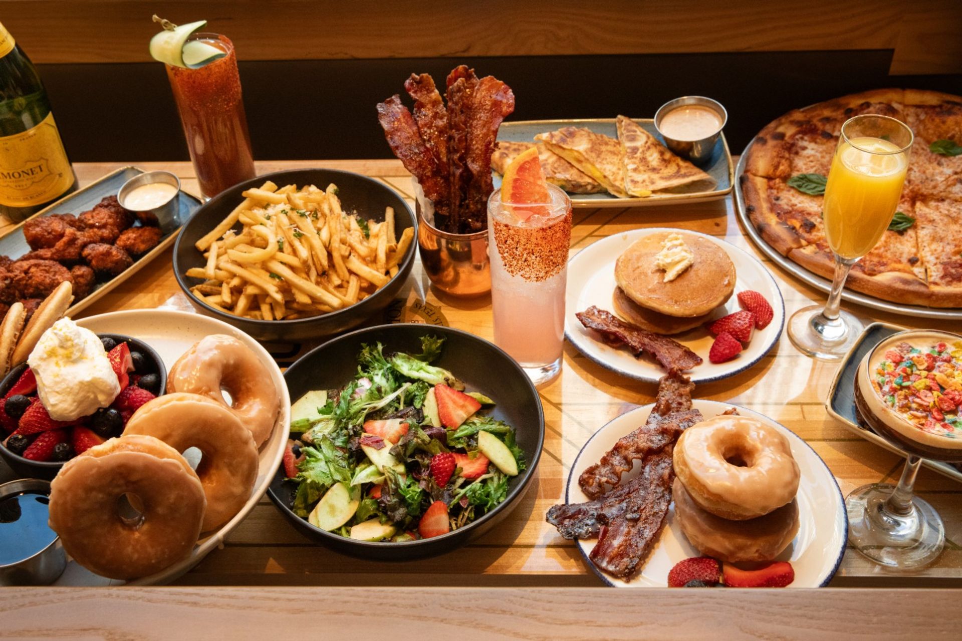Vibrant brunch spread on a wooden table: pancakes with bacon and strawberries, glazed donuts, crispy bacon strips, fries, mixed green salad, fried chicken bites, pizza, quesadilla and colorful cocktails.
