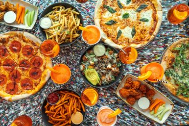 Top-down view of a casual pizzeria spread with pepperoni, margherita and arugula pizzas, fries and sweet potato fries, buffalo wings, avocado salad, and orange cocktails on a colorful table.