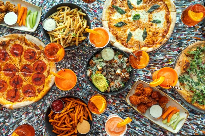 Top-down view of a casual pizzeria spread with pepperoni, margherita and arugula pizzas, fries and sweet potato fries, buffalo wings, avocado salad, and orange cocktails on a colorful table.