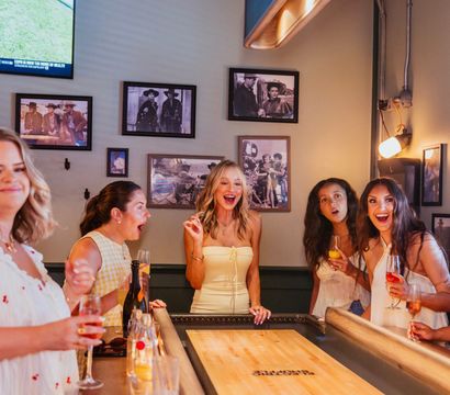 Five women laughing and cheering around a shuffleboard table in a cozy indoor bar, holding cocktails and wine beneath vintage framed photos.