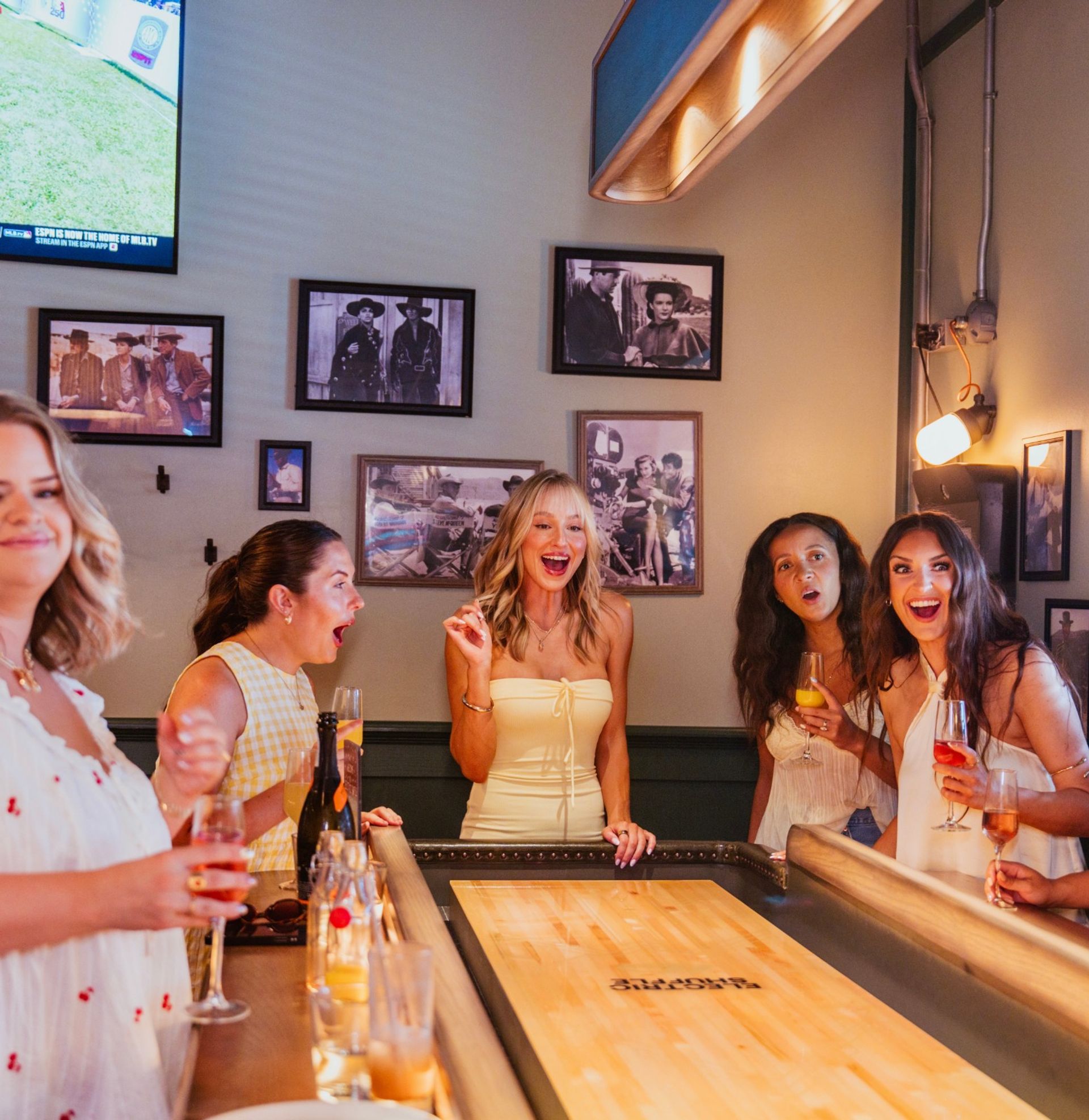 Five women laughing and cheering around a shuffleboard table in a cozy indoor bar, holding cocktails and wine beneath vintage framed photos.