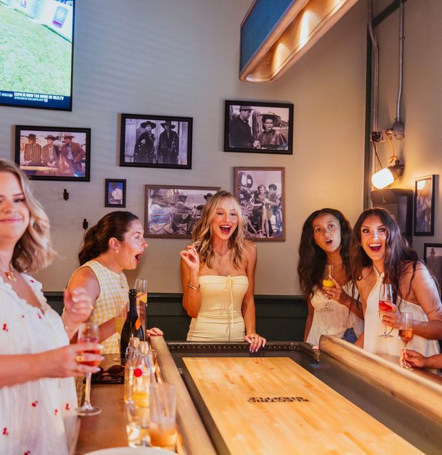 Five women laughing and cheering around a shuffleboard table in a cozy indoor bar, holding cocktails and wine beneath vintage framed photos.