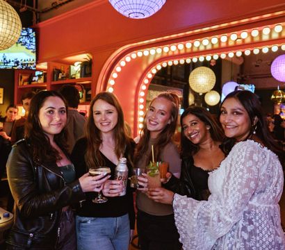 Group of five friends smiling and holding cocktails at a lively bar with globe pendant lights, pink marquee mirror lights and colorful nightlife ambiance
