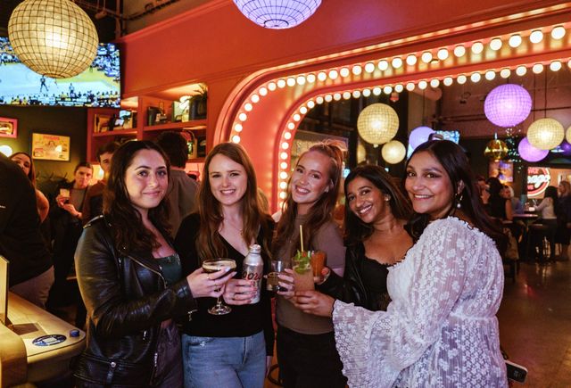 Group of five friends smiling and holding cocktails at a lively bar with globe pendant lights, pink marquee mirror lights and colorful nightlife ambiance