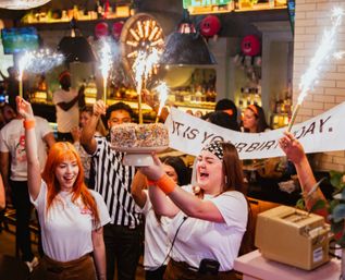 Lively bar/restaurant birthday party with staff carrying a sprinkle-covered cake topped with lit sparklers while a cheering crowd holds a banner under warm pendant lights