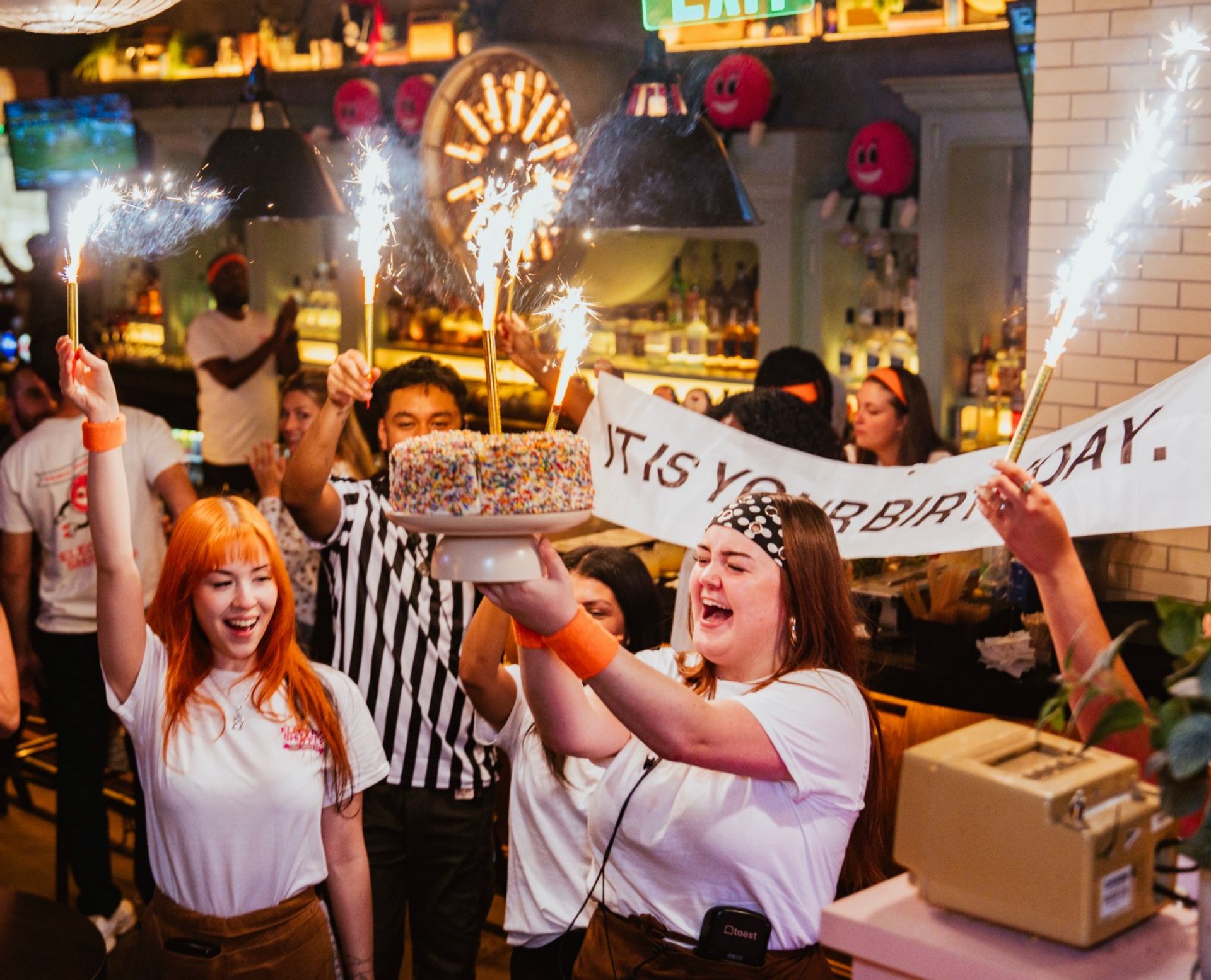 Lively bar/restaurant birthday party with staff carrying a sprinkle-covered cake topped with lit sparklers while a cheering crowd holds a banner under warm pendant lights