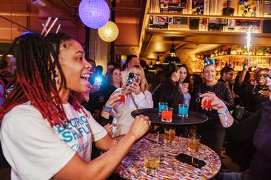 Bartender handing a tray of bright red and blue cocktail shots to cheering guests in a lively indoor bar party with people taking photos
