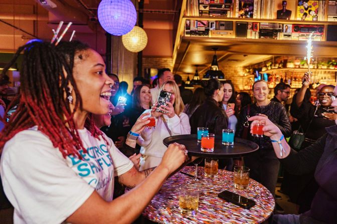 Bartender handing a tray of bright red and blue cocktail shots to cheering guests in a lively indoor bar party with people taking photos