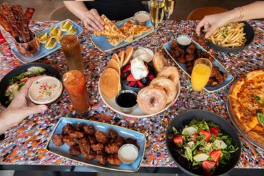 Vibrant brunch spread on a colorful table: glazed donuts and mini pancakes with berries, pizza, fries, fried chicken bites, salads, deviled eggs, crispy bacon, cocktails and mimosas with hands reaching in.