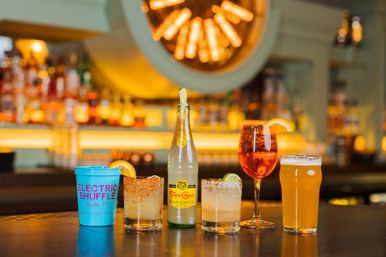 Colorful drinks lined up on a bar counter: a bright blue plastic cup, two salt-rimmed citrus cocktails, a sparkling water bottle, an orange aperitif in a wine glass with an orange slice, and a pint of beer against warm, backlit liquor shelves.