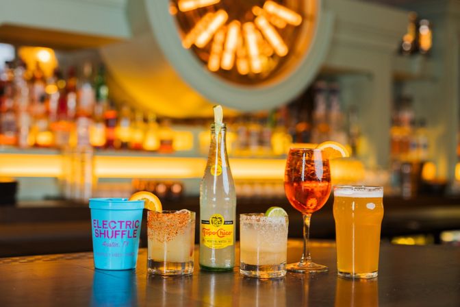 Colorful drinks lined up on a bar counter: a bright blue plastic cup, two salt-rimmed citrus cocktails, a sparkling water bottle, an orange aperitif in a wine glass with an orange slice, and a pint of beer against warm, backlit liquor shelves.
