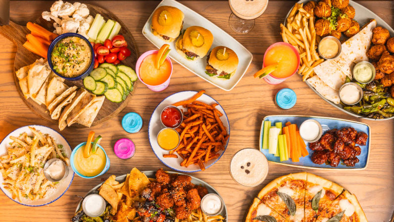 Flat-lay of a colorful casual dining spread with pizza, sliders, chicken wings, sweet potato fries, fries, quesadillas, veggie-and-pita dip platter, chips, dipping sauces and frozen cocktails on a wooden table