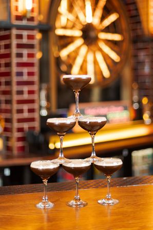 Six espresso martinis stacked like a cocktail tower on a wooden bar counter, creamy foam and warm golden bokeh lights in a cozy bar interior