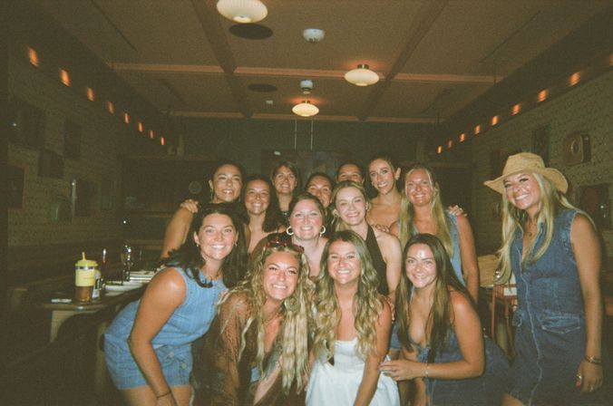 Group photo of smiling women posing together inside a dimly lit restaurant or bar, casual summer outfits, denim and a cowboy hat