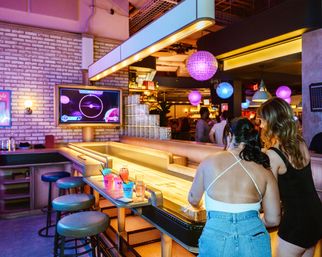 Two women leaning over a long shuffleboard table in a neon-lit urban bar with purple hanging lanterns, a wall-mounted TV, round stools, and colorful cocktails on the counter.