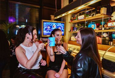 Three friends toasting cocktails at a lively retro-style bar counter with colorful shelves and a neon-lit TV in the background — cheerful night out scene