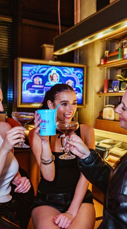 Three friends toasting cocktails at a lively retro-style bar counter with colorful shelves and a neon-lit TV in the background — cheerful night out scene