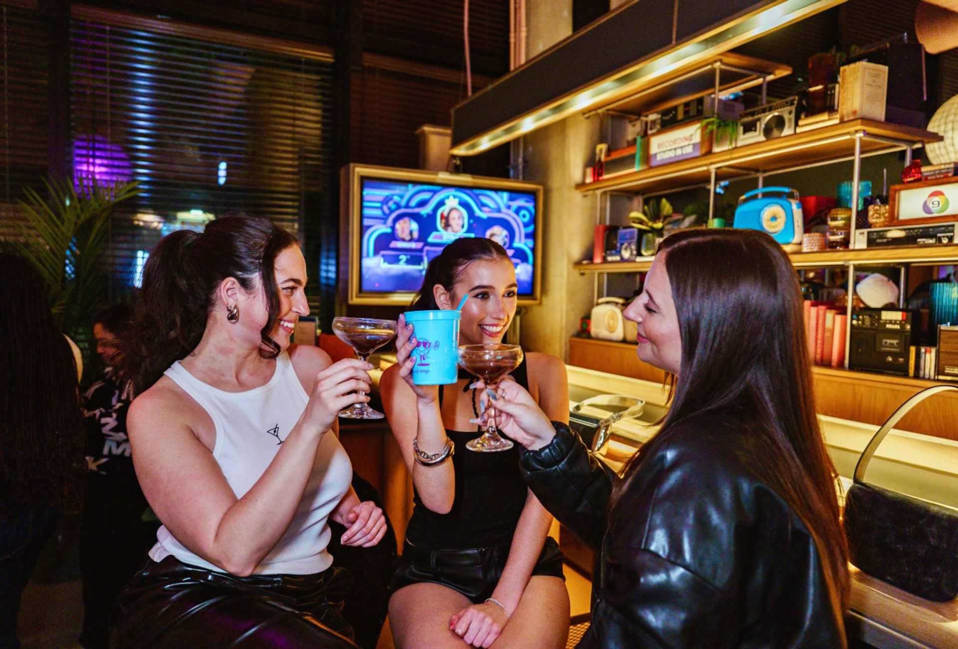 Three friends toasting cocktails at a lively retro-style bar counter with colorful shelves and a neon-lit TV in the background — cheerful night out scene