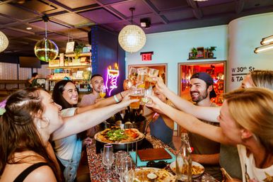 Friends clinking glasses—beers, cocktails and pizza—at a colorful, trendy bar with disco balls, neon lights and a lively celebration atmosphere