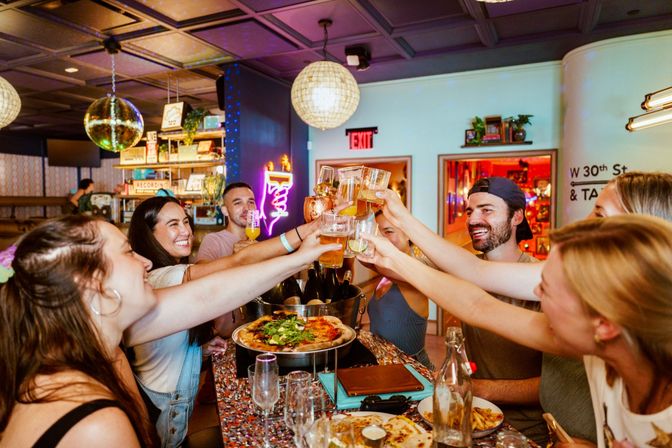 Friends clinking glasses—beers, cocktails and pizza—at a colorful, trendy bar with disco balls, neon lights and a lively celebration atmosphere