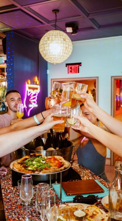 Friends clinking glasses—beers, cocktails and pizza—at a colorful, trendy bar with disco balls, neon lights and a lively celebration atmosphere