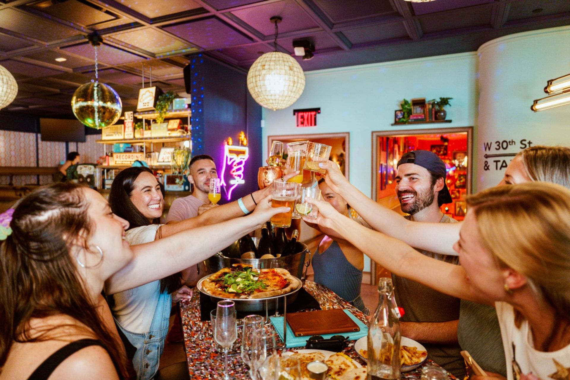Friends clinking glasses—beers, cocktails and pizza—at a colorful, trendy bar with disco balls, neon lights and a lively celebration atmosphere