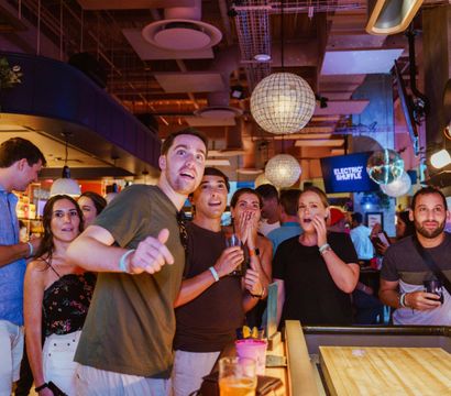 Group of friends at a lively indoor bar playing shuffleboard, cheering and reacting to a shot beneath hanging globe lights with colorful drinks on the counter — upbeat nightlife scene.