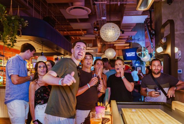 Group of friends at a lively indoor bar playing shuffleboard, cheering and reacting to a shot beneath hanging globe lights with colorful drinks on the counter — upbeat nightlife scene.