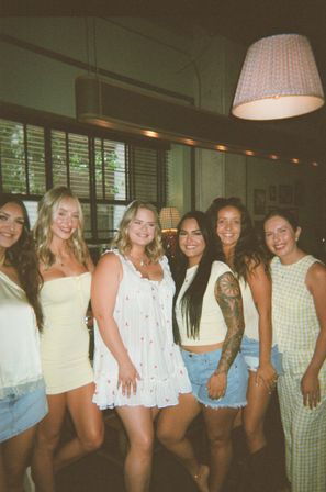 Six friends in summer outfits smiling and posing inside a cozy café under warm pendant lighting and window blinds.