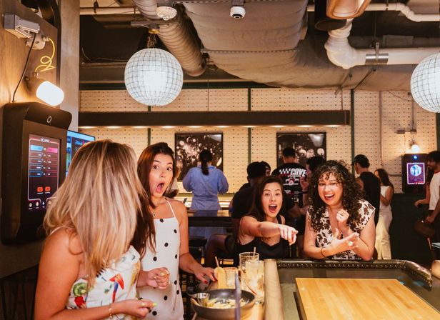 Four women laughing and reacting around a shuffleboard table at an indoor game bar, drinks on the counter, globe pendant lights and exposed ducts in a lively urban nightlife setting.