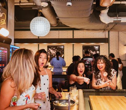 Four women laughing and reacting around a shuffleboard table at an indoor game bar, drinks on the counter, globe pendant lights and exposed ducts in a lively urban nightlife setting.