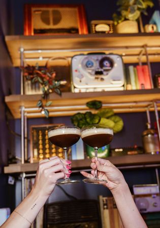 Two espresso martini cocktails clinking in coupe glasses in a cozy home-bar setting with wooden shelves, potted plants and vintage audio gear