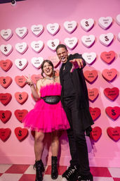 Two friends posing at a pink Valentine-themed photo wall covered in candy-heart messages; woman in hot-pink tulle dress flashes a peace sign, man in black points and laughs.