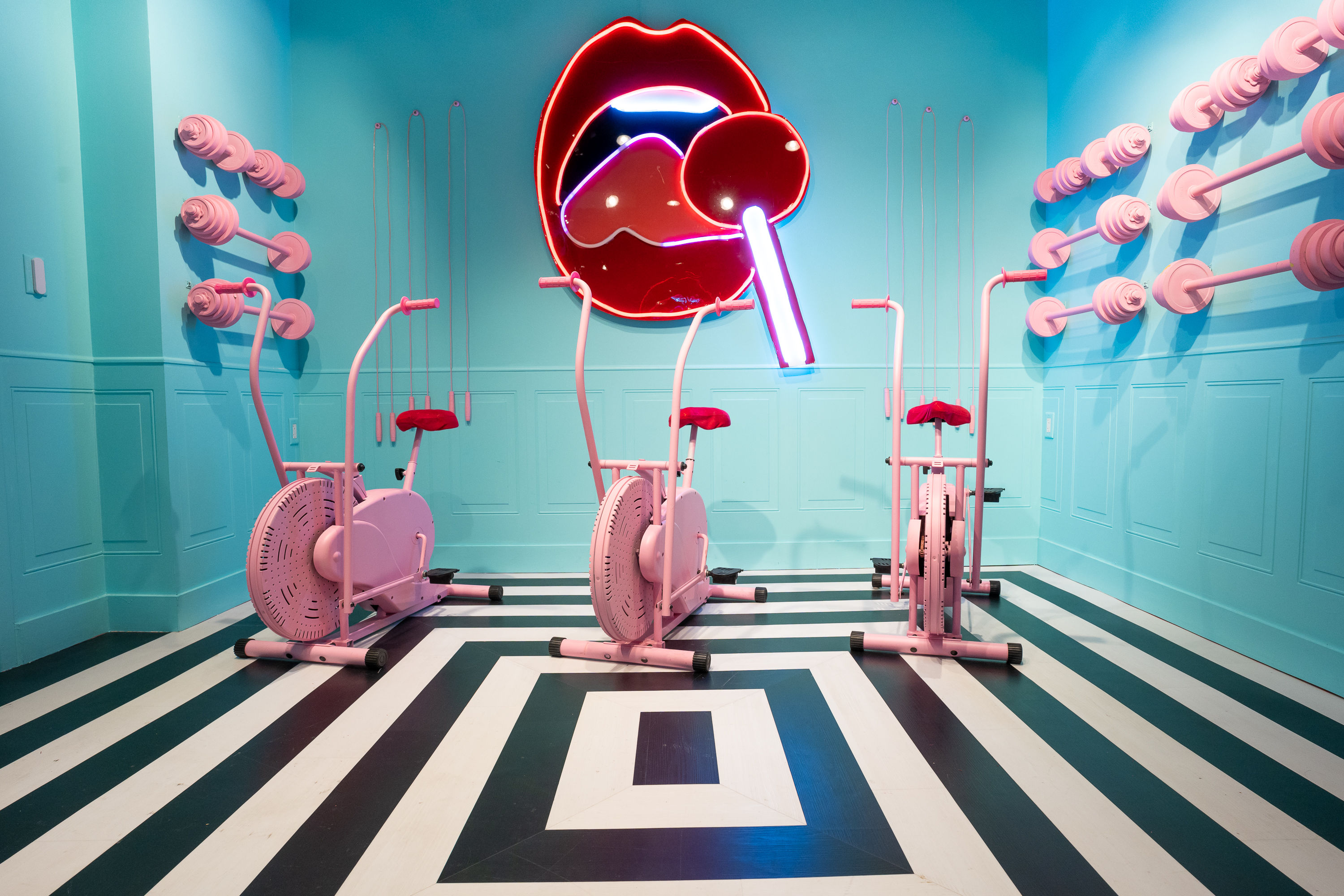 Three pink exercise bikes in a turquoise indoor fitness studio with black-and-white striped floor, neon red lips lollipop wall sign and pink weight racks — playful retro workout room.