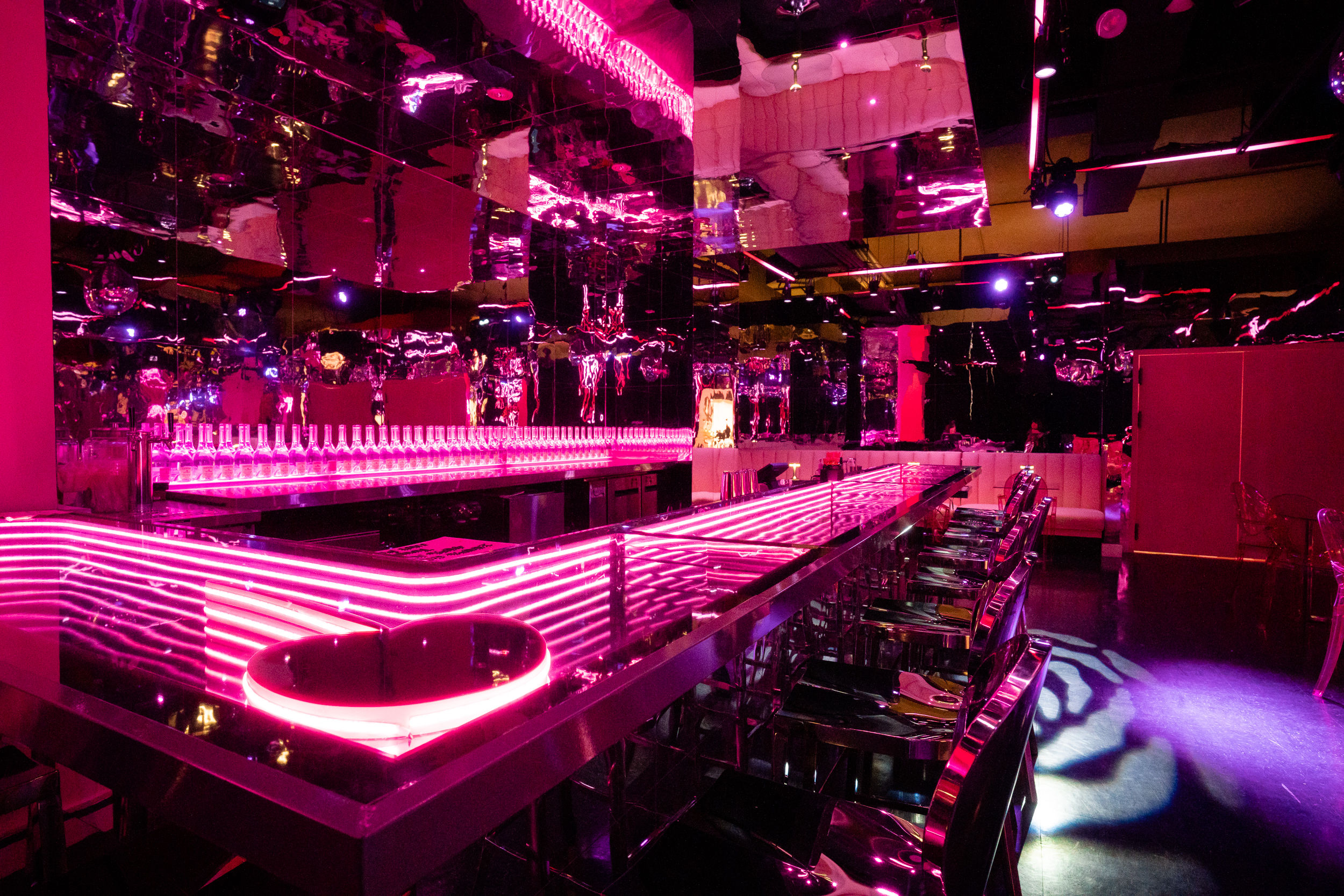 Neon-pink nightclub interior with mirrored walls, long LED-lit bar counter, rows of translucent bar stools and backlit bottles creating a futuristic lounge vibe
