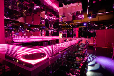 Neon-pink nightclub interior with mirrored walls, long LED-lit bar counter, rows of translucent bar stools and backlit bottles creating a futuristic lounge vibe