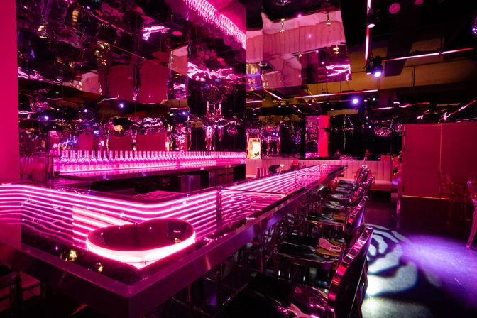 Neon-pink nightclub interior with mirrored walls, long LED-lit bar counter, rows of translucent bar stools and backlit bottles creating a futuristic lounge vibe