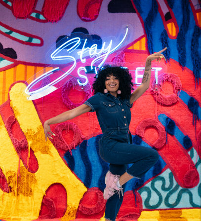 Smiling woman with curly hair in a denim jumpsuit and pink sneakers mid-jump in front of a vibrant street-art mural and neon "Stay Sweet" sign