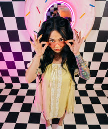 Young woman in a yellow lace-trim dress adjusting pink sunglasses on a black-and-white checkered floor, neon donut wall art with colorful sprinkles and a vibrant sleeve tattoo in a retro pastel studio.