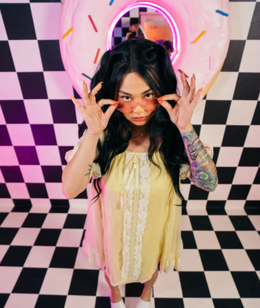 Young woman in a yellow lace-trim dress adjusting pink sunglasses on a black-and-white checkered floor, neon donut wall art with colorful sprinkles and a vibrant sleeve tattoo in a retro pastel studio.